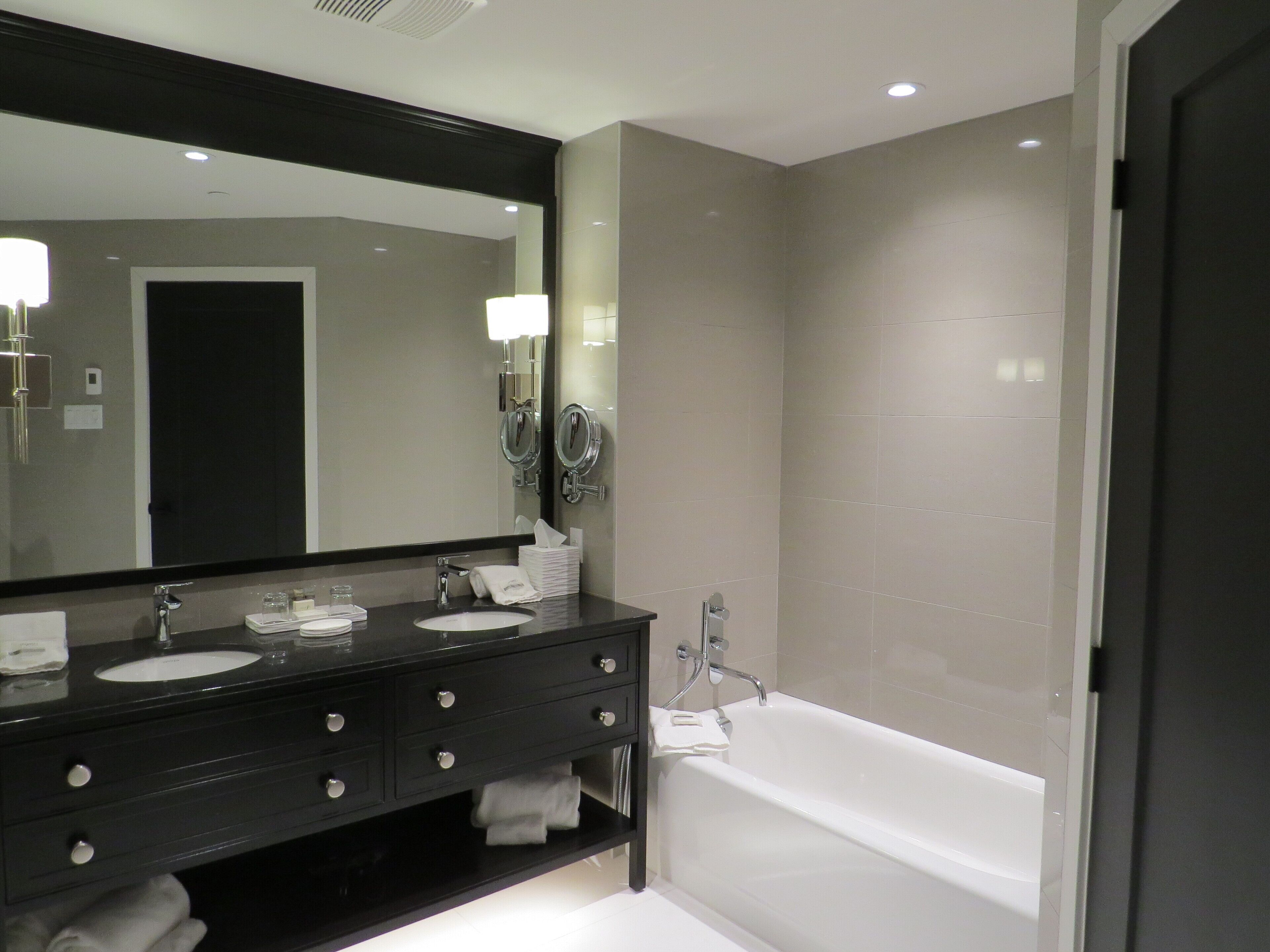 A modern bathroom with a black vanity, two sinks, a large mirror, and a white bathtub.