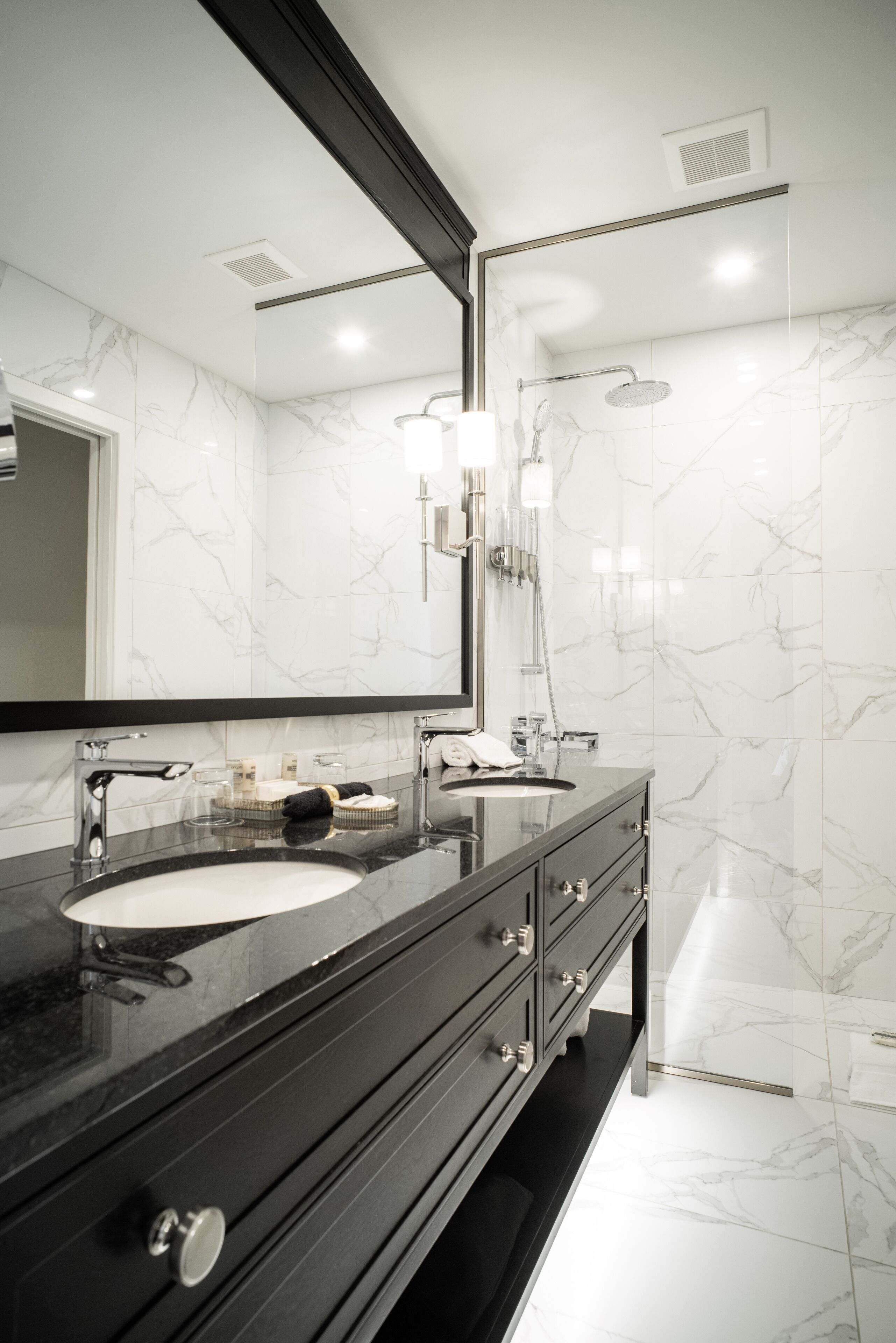 Un baño moderno con tocador negro, azulejos de mármol blanco, un espejo grande y una zona de ducha.