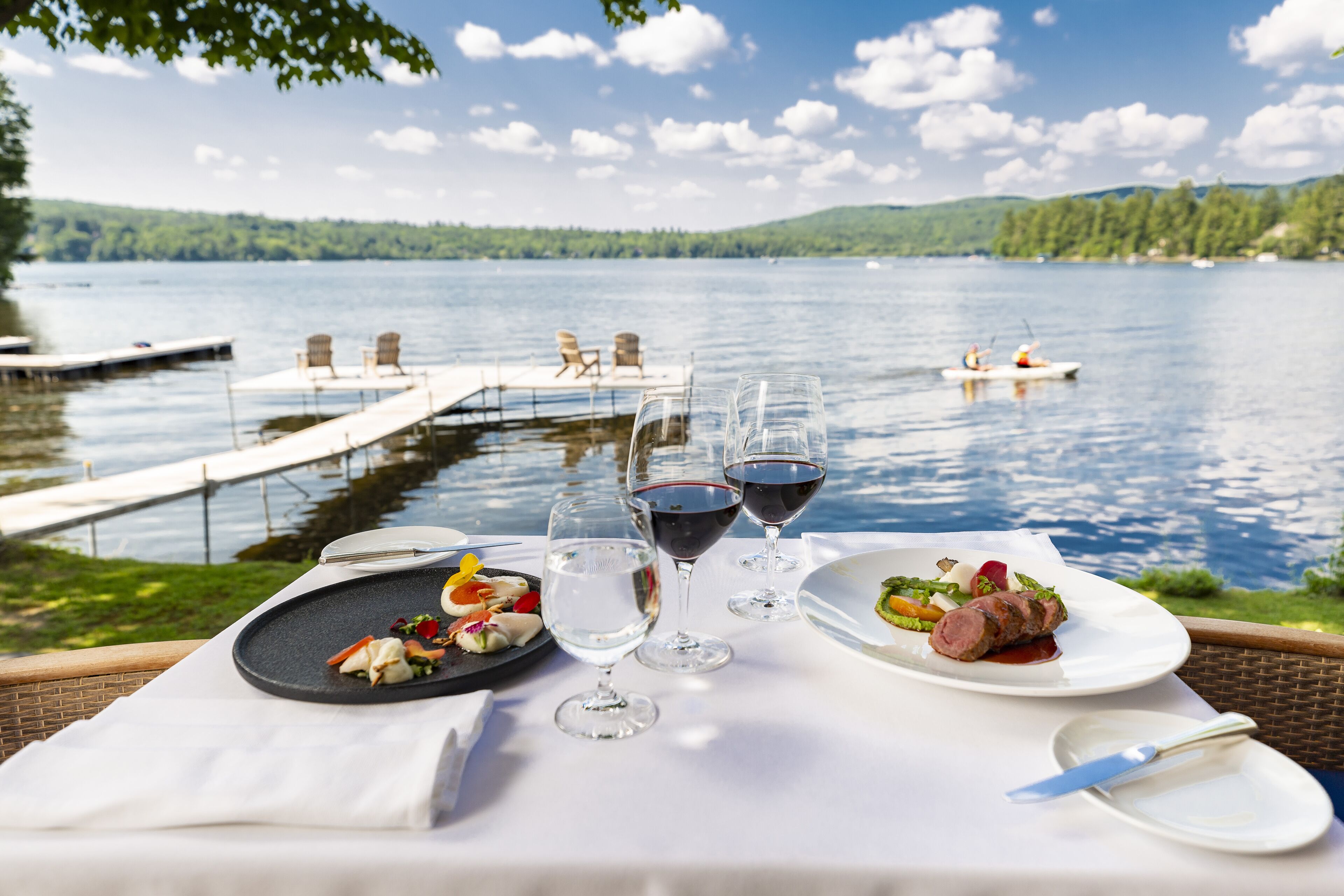 A dining table with plates of food and wine glasses, overlooking a lake with a dock and chairs.