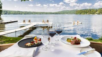 A dining table with plates of food and wine glasses, overlooking a lake with a dock and chairs.