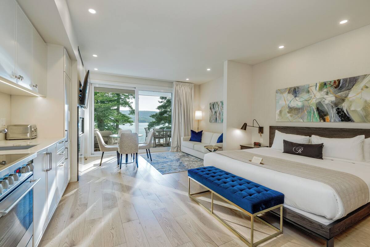 A modern hotel room with a large bed, a dining area, and a view of the outdoors.