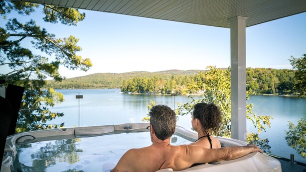 A couple in a hot tub overlooking a lake with trees and hills in the background.