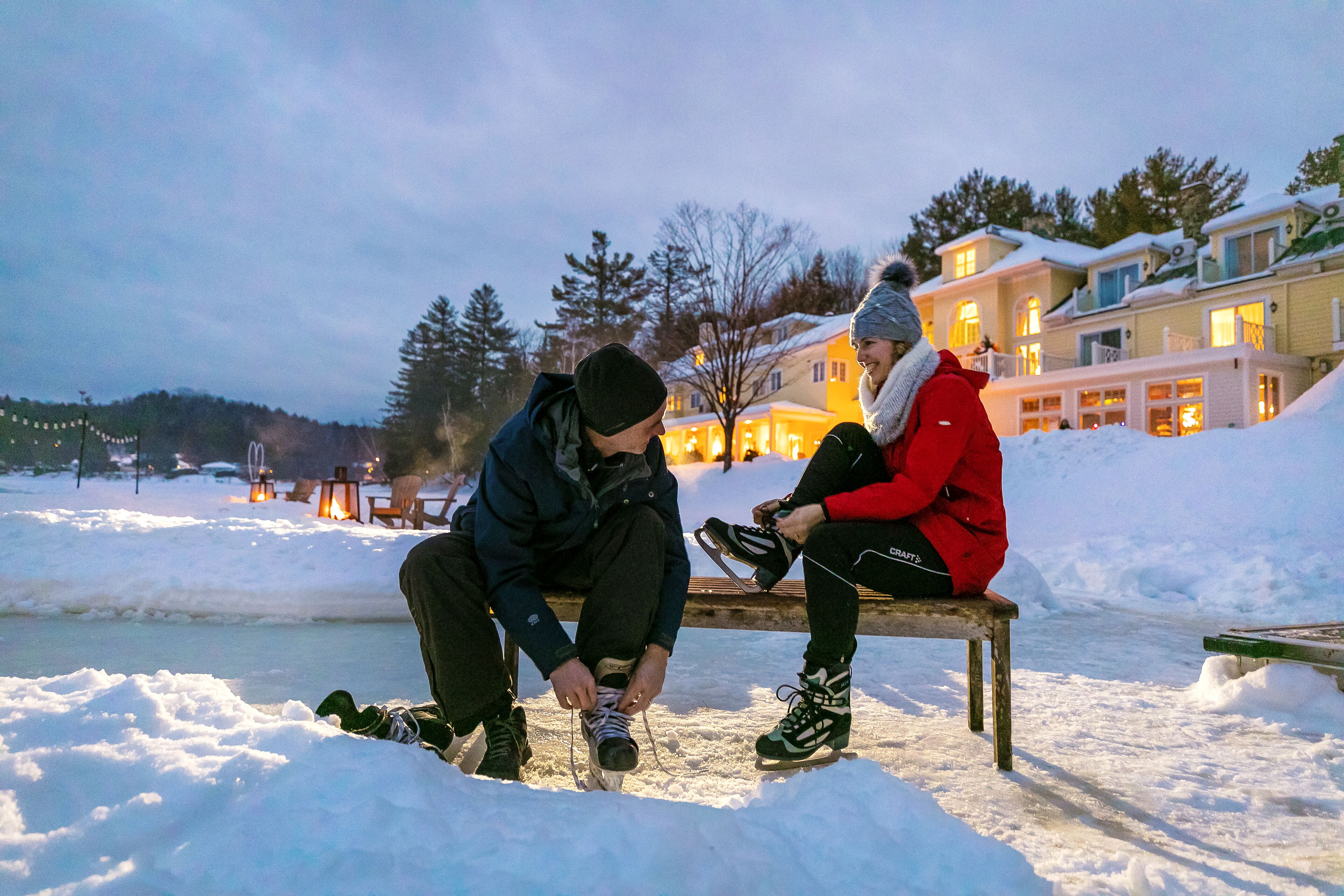 Two people sitting on a bench in a snowy outdoor area with a body of water and buildings in the background.