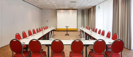 Een vergaderzaal met een lange tafel, rode stoelen en een vloerbedekking.
