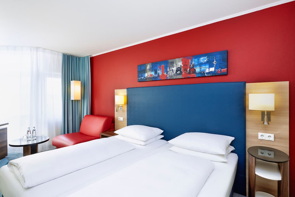 A hotel room with a large bed, a red armchair, a small table with a lamp, and a painting on the wall.