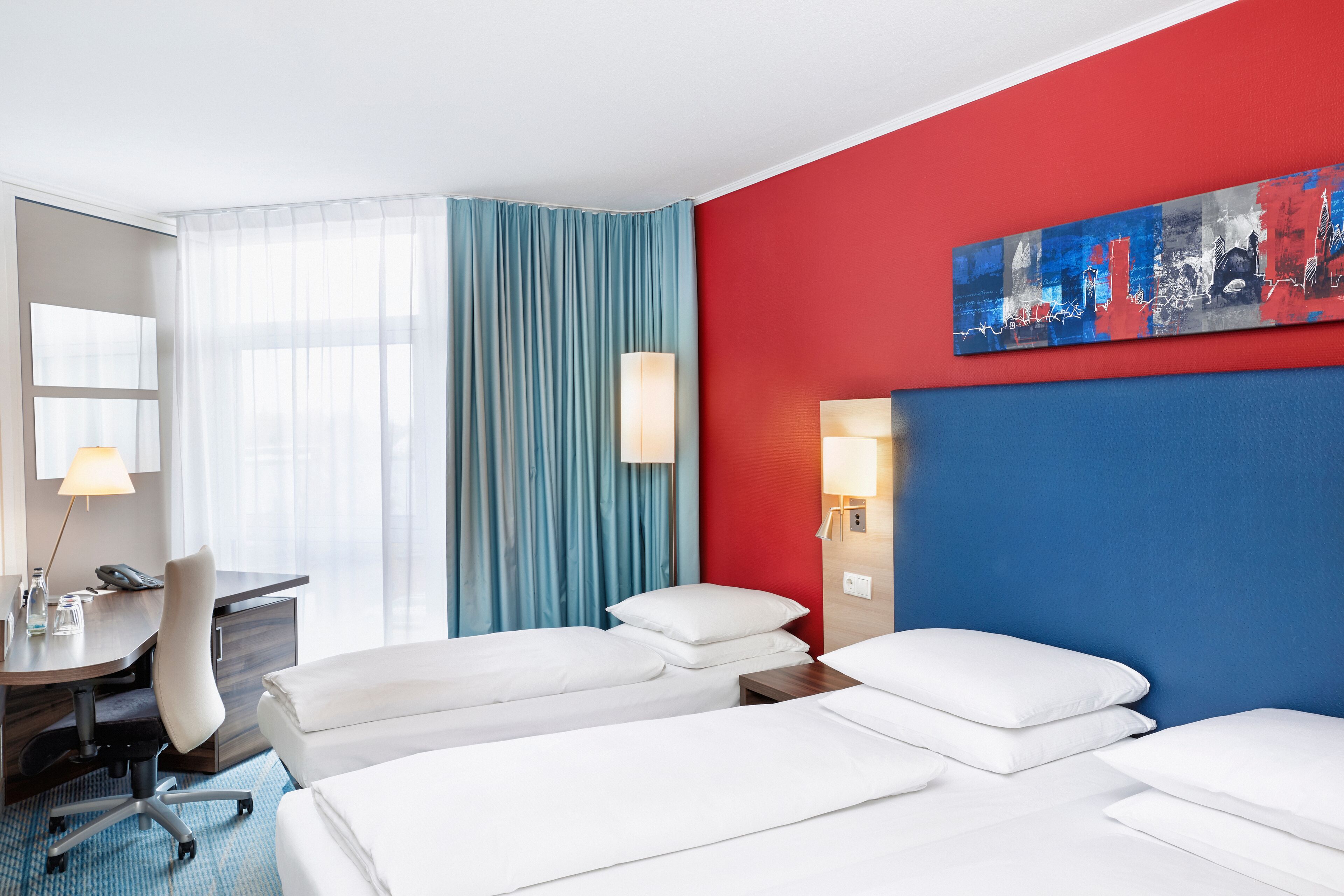 Une chambre d’hôtel avec un mur d’accent rouge, une grande fenêtre avec des rideaux bleus, un bureau avec une lampe et deux lits avec du linge de lit blanc.