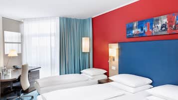 A hotel room with a red accent wall, a large window with blue curtains, a desk with a lamp, and two beds with white linens.