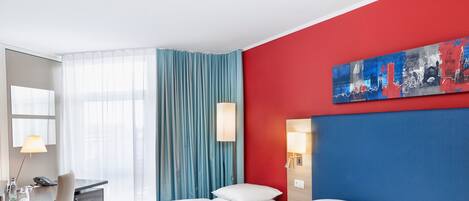 Habitación de hotel con una pared de acento rojo, un ventanal con cortinas azules, un escritorio con lámpara y dos camas con sábanas blancas.