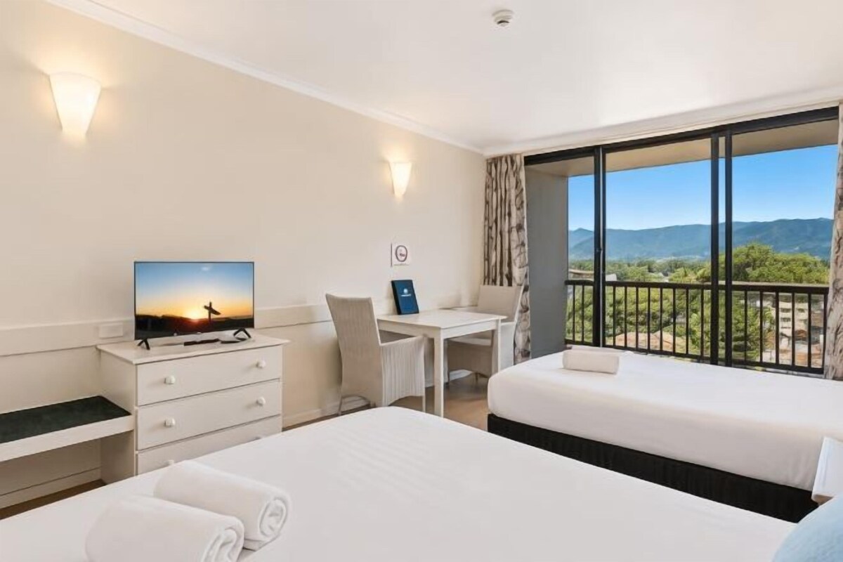 A hotel room with two beds, a desk, a TV, and a balcony with a view.