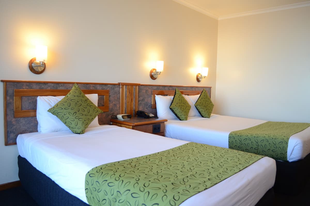 A hotel room with two beds, each with a green patterned bedspread, white linens, and matching green pillows. There are bedside tables with lamps and a telephone.