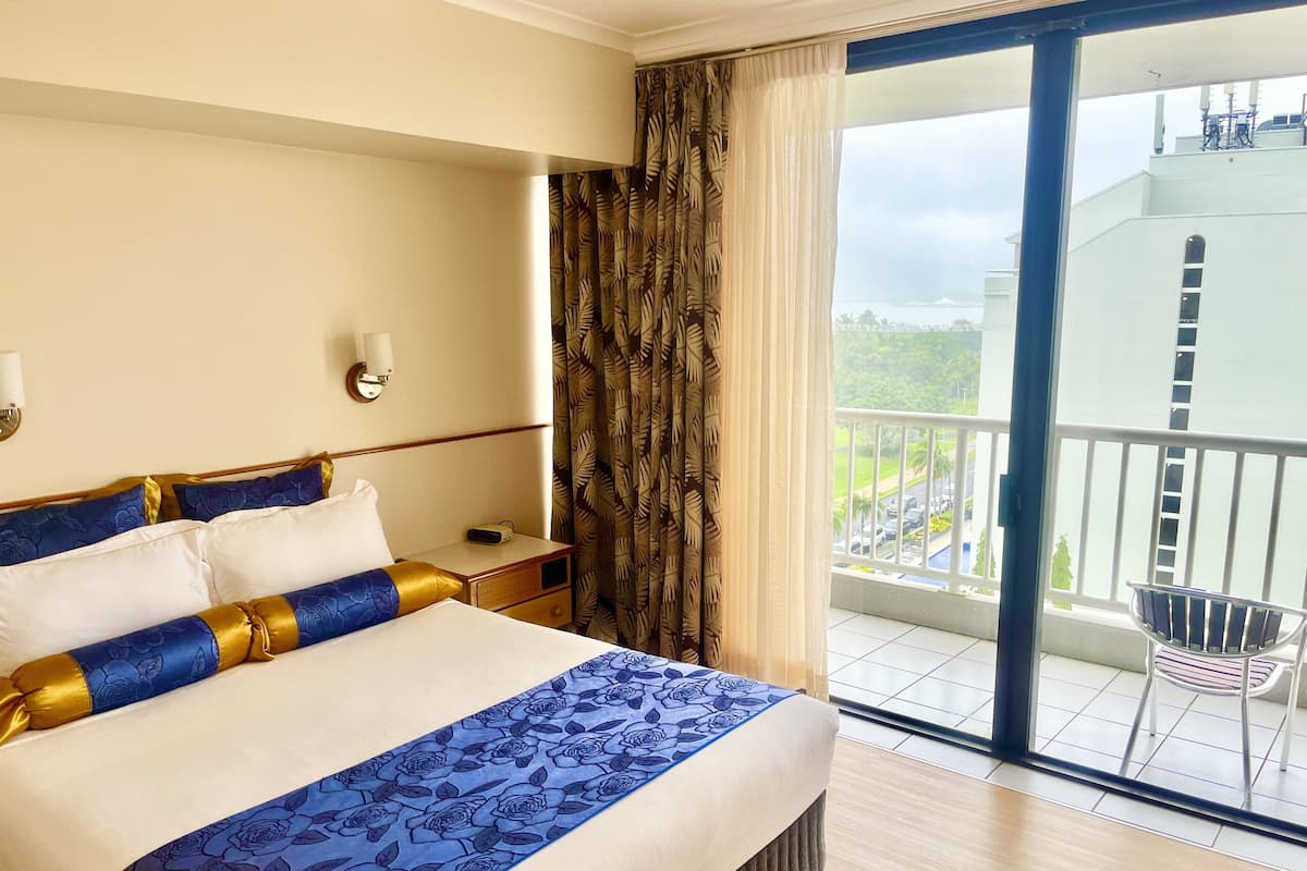 A hotel room with a large bed, a nightstand, a wall-mounted lamp, and a view of a balcony with a railing and a chair.