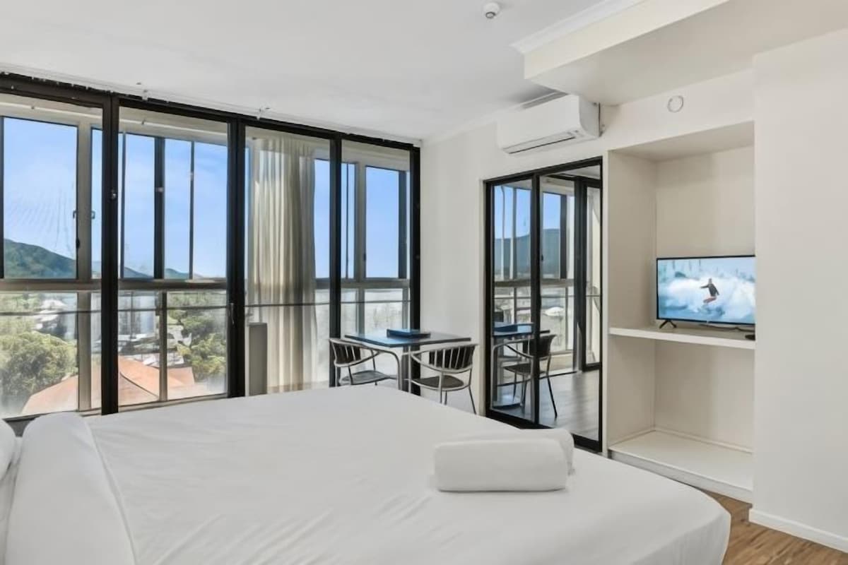 A hotel room with a large bed, a television, a balcony with a table and chairs, and a view of the outside.