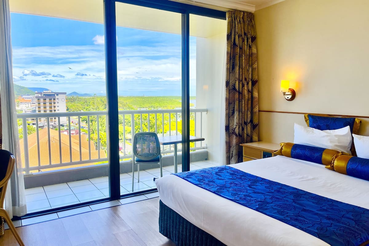 A hotel room with a large bed, a balcony with a view of buildings and greenery, a chair, and a nightstand.
