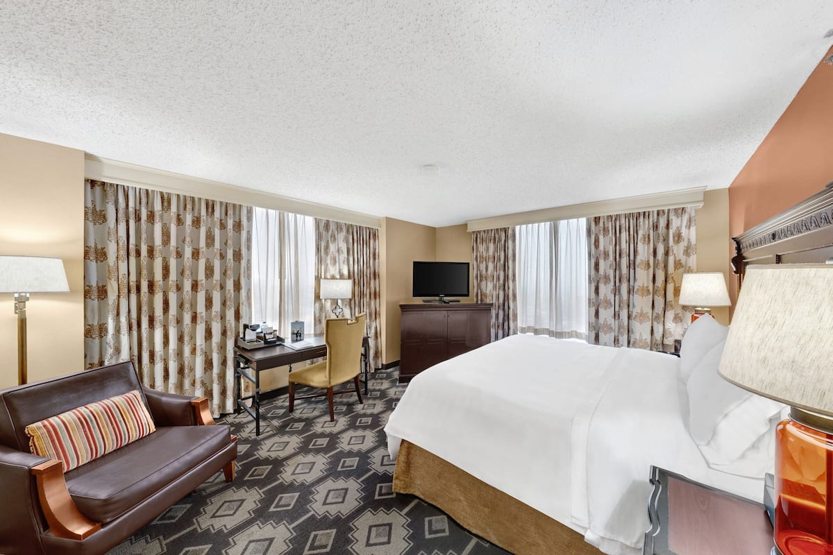 A hotel room with a large bed, a desk with a chair, a television, and curtains with a pattern.