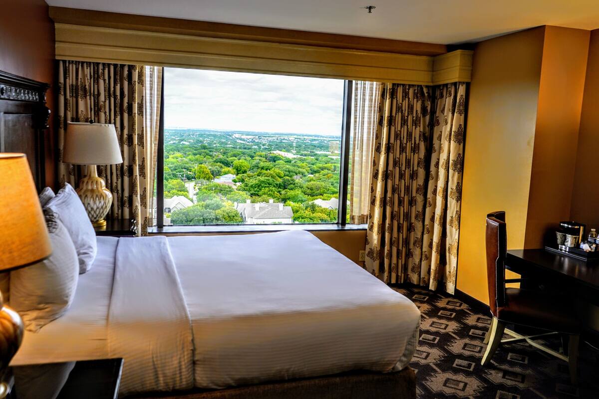 A hotel room with a large bed, a desk, a lamp, and a view of a green landscape through the window.
