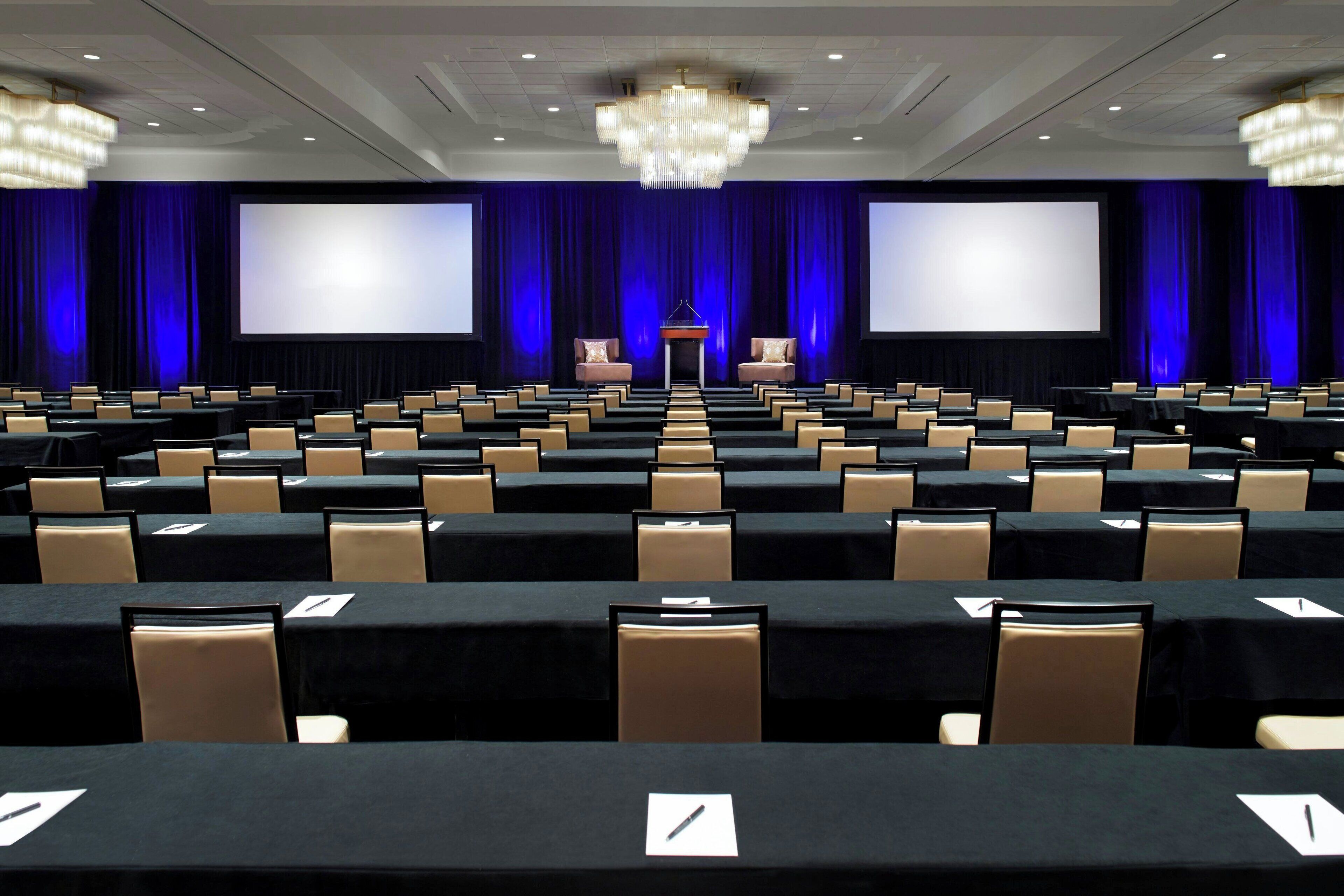 A conference room with rows of tables and chairs, a large screen, and a podium.