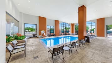 Uma área à beira da piscina com cadeiras e mesas, piscina e grandes janelas com vista para o exterior.