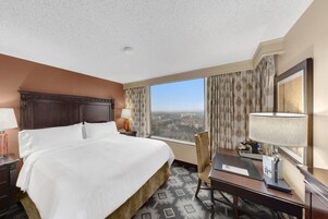 A hotel room with a large bed, a desk, a chair, a lamp, and a view of the city.