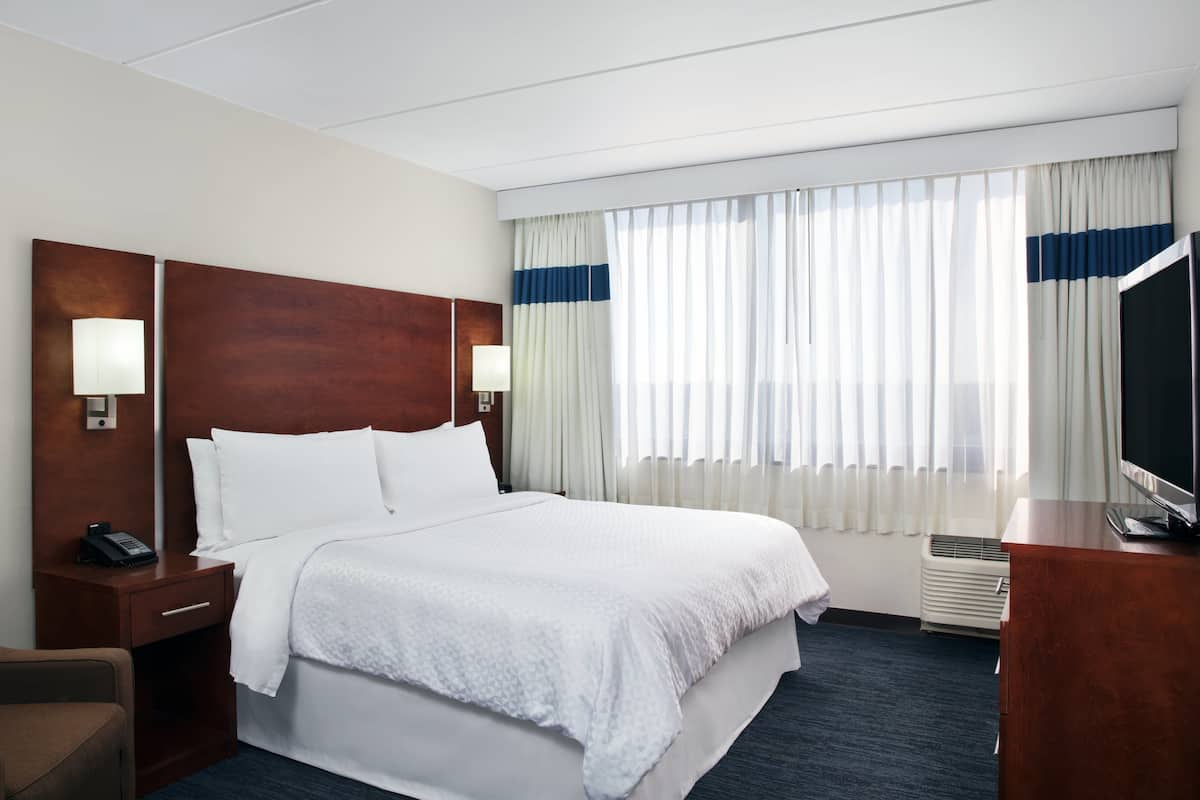 A hotel room with a large bed, a wooden headboard, two bedside tables with lamps, a TV on a wooden dresser, and a window with sheer curtains.