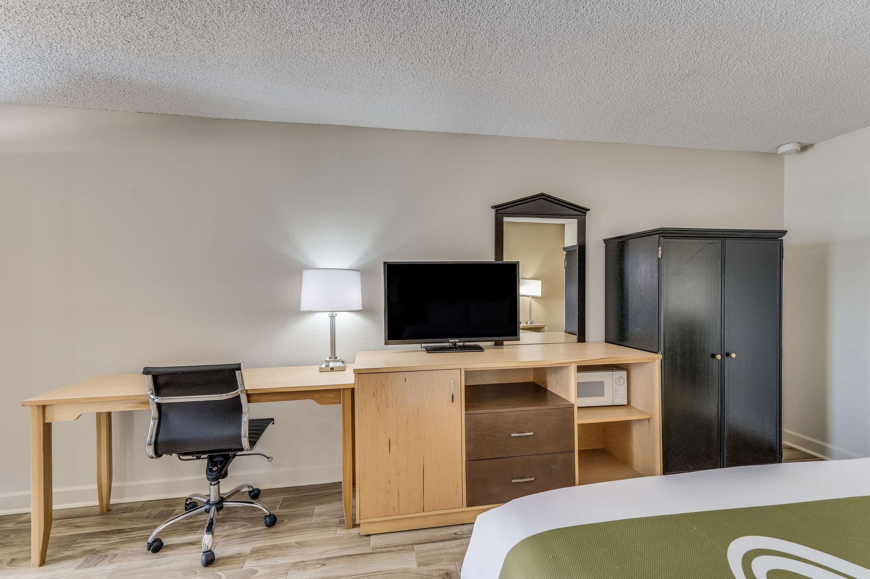Une chambre d’hôtel avec un bureau en bois, un fauteuil de bureau noir, une télévision, une armoire et un lit recouvert d’une couverture verte.