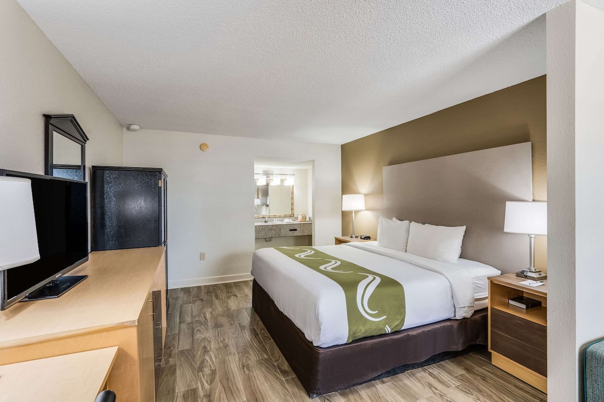 A hotel room with a bed, bedside table, lamp, TV, and a mirror.