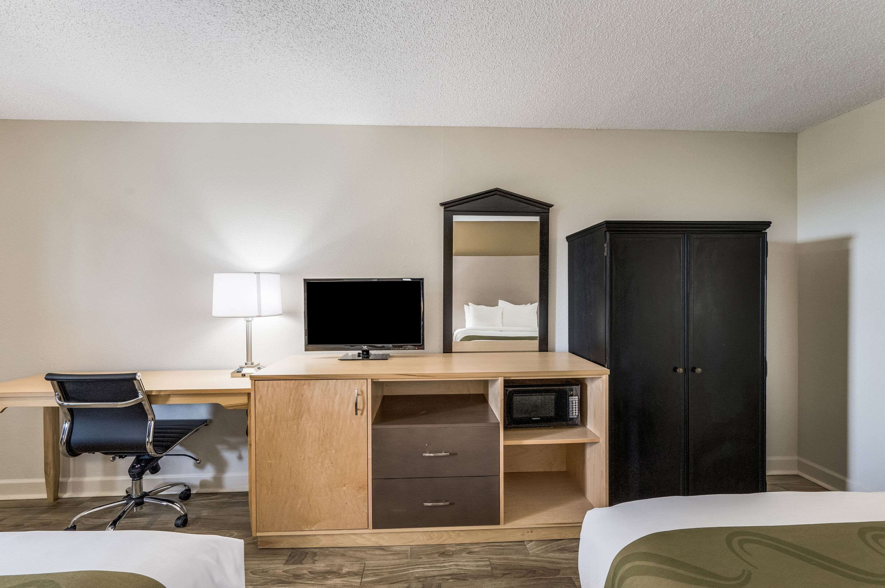 A hotel room with a wooden desk, a black office chair, a flat-screen TV, a mirror, a wardrobe, and two beds.