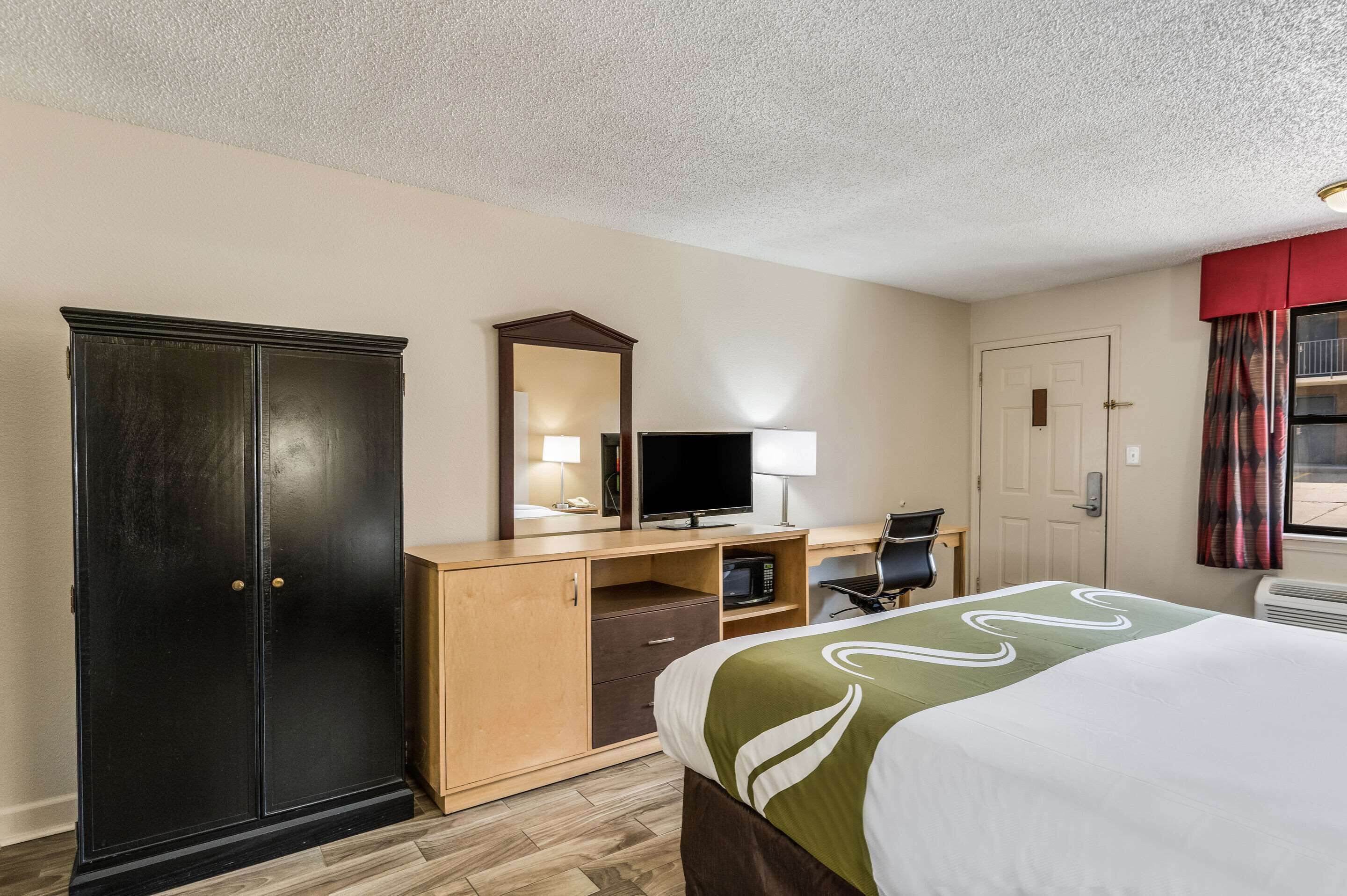 A hotel room with a bed, a desk with a computer, a wardrobe, a mirror, and a door.