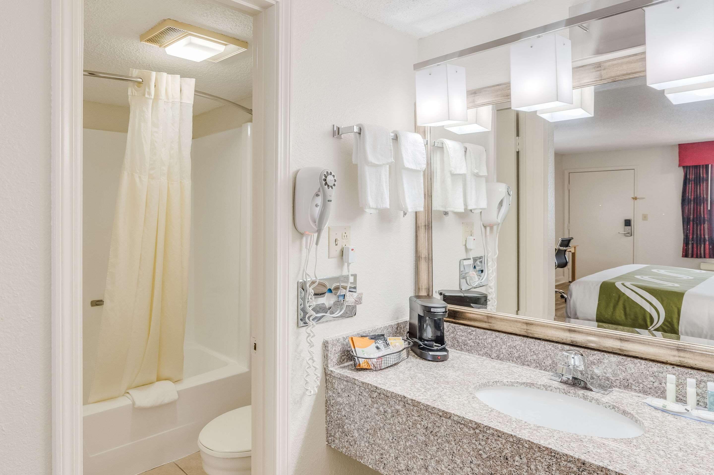 A bathroom with a shower, toilet, sink, and a mirror reflecting a bed with a green bedspread.