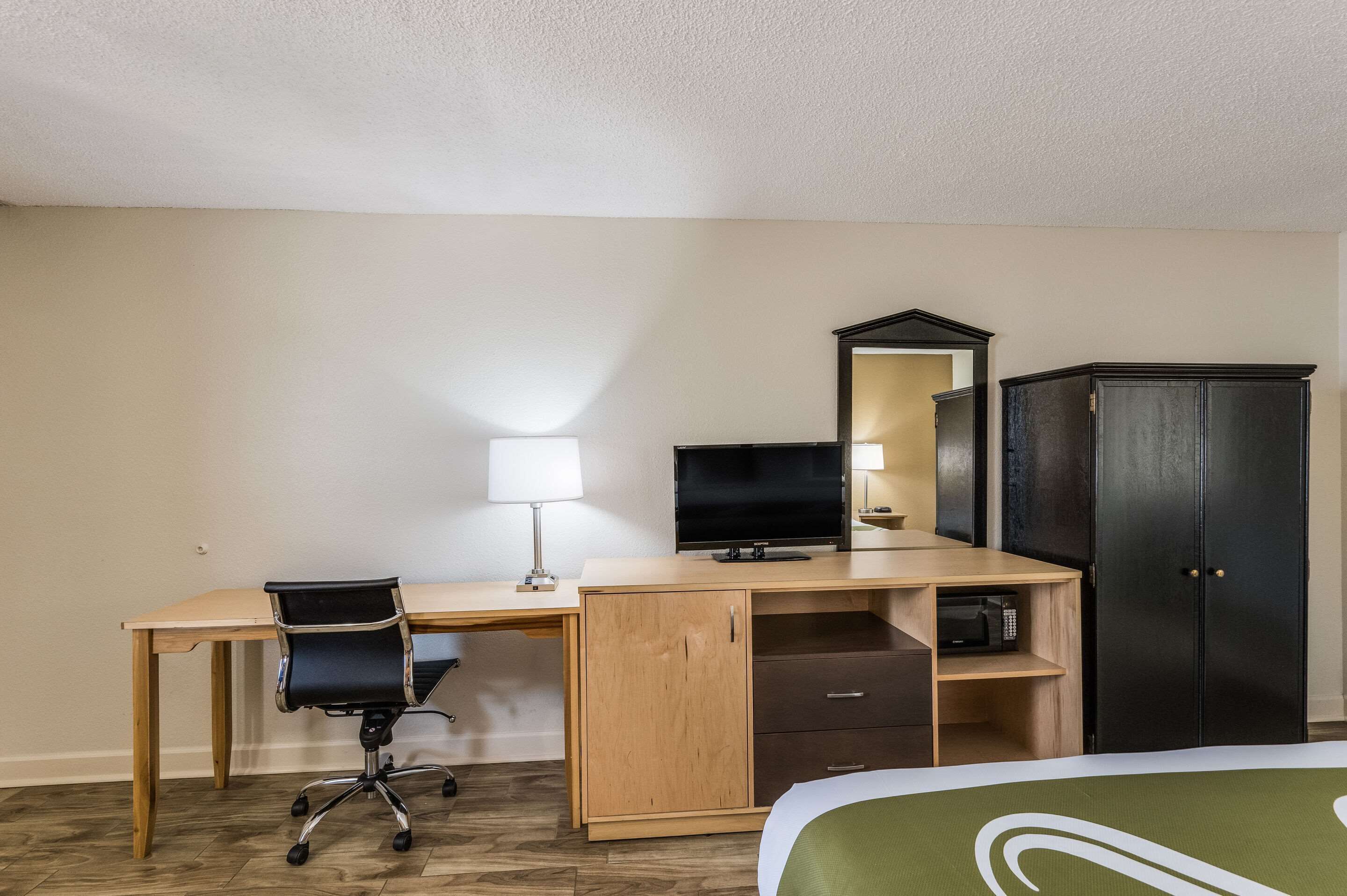 A hotel room with a wooden desk, a black office chair, a television, a wardrobe, and a bed with a green and white bedspread.