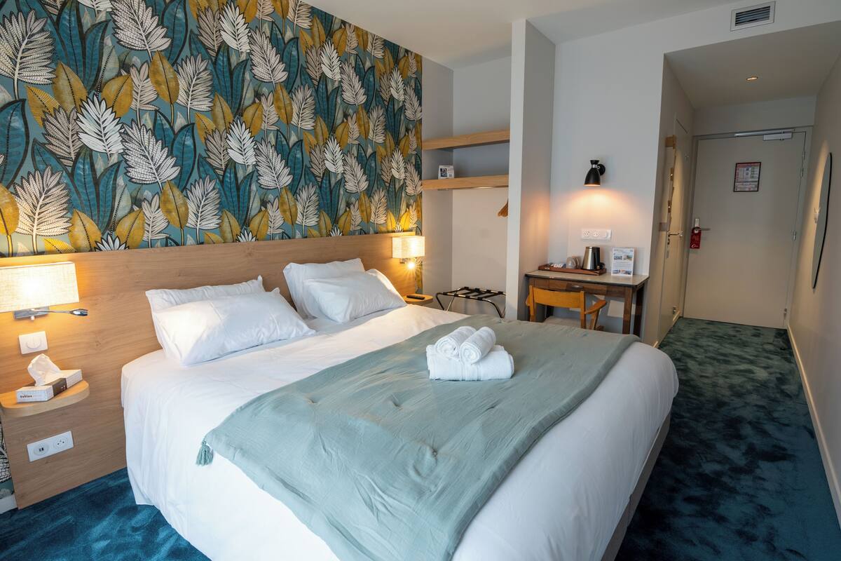 Ein Hotelzimmer mit einem Bett, einem Nachttisch, einer Lampe und einer Wand mit Blätermotiv.