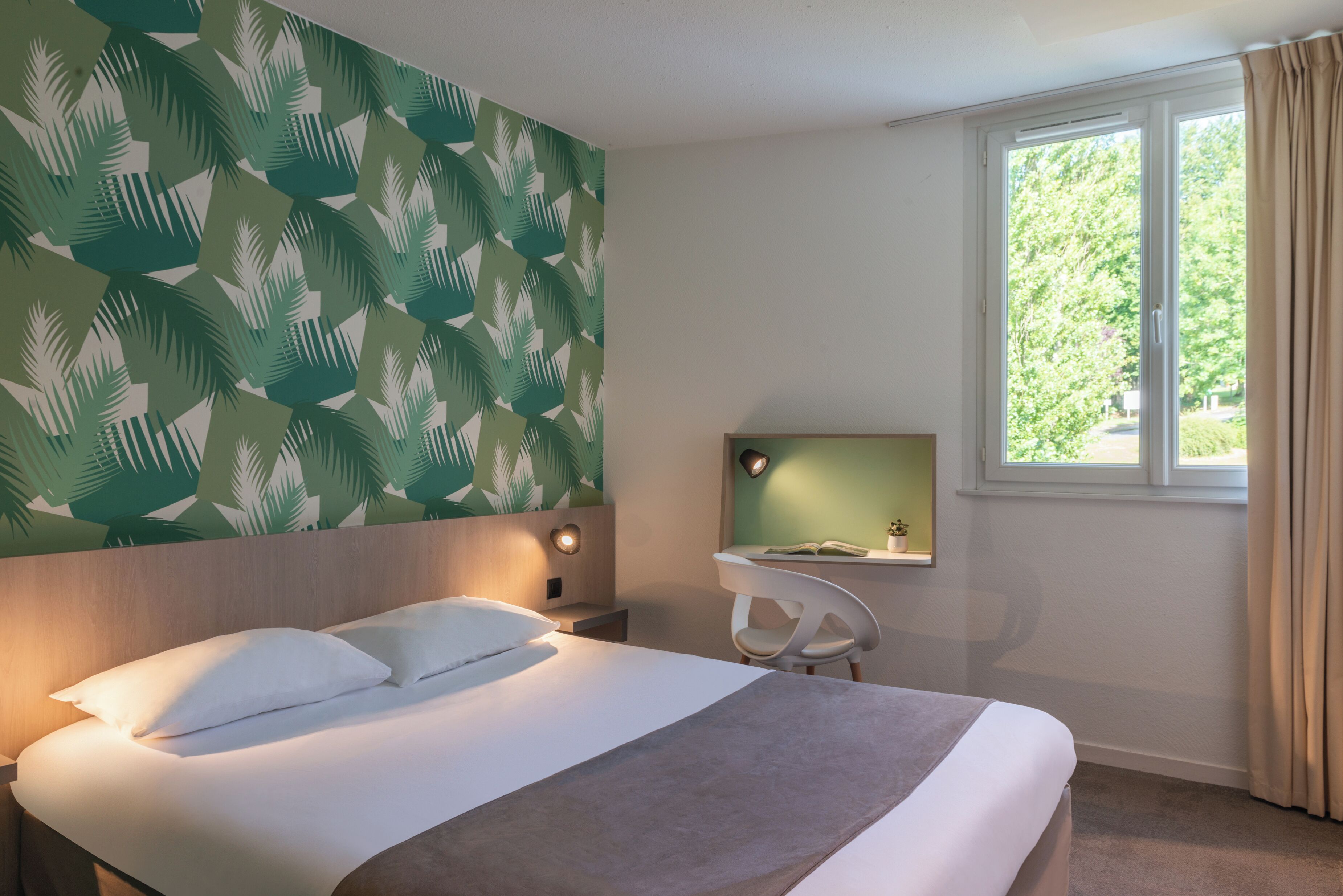 Habitación de hotel con cama, escritorio con silla, ventana con cortinas y pared con diseño de hojas.