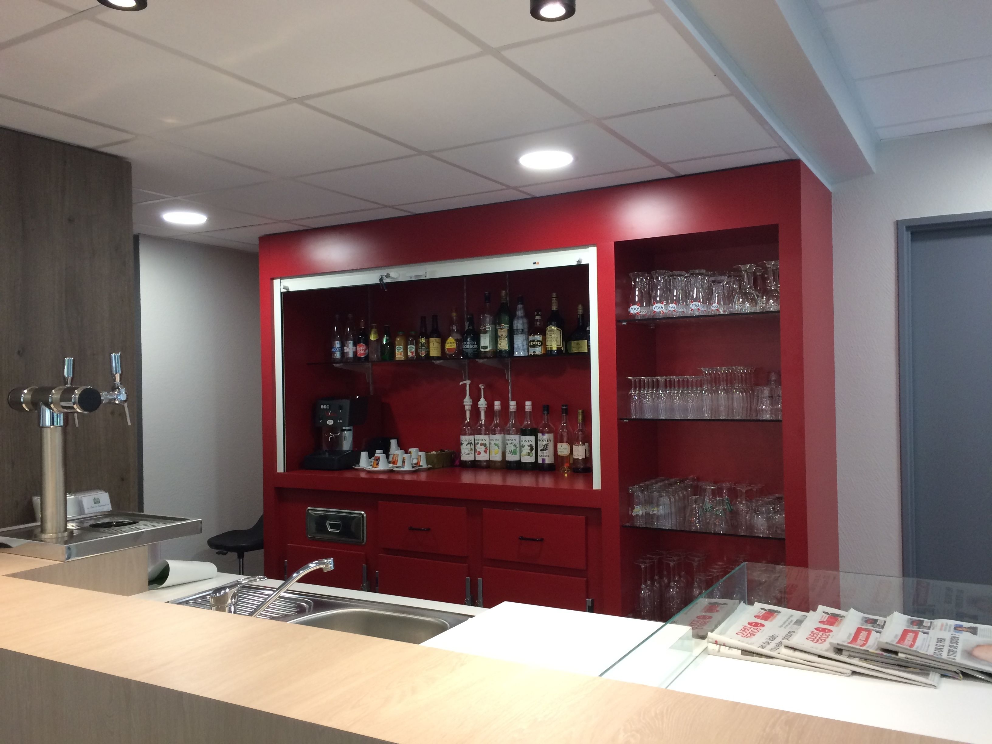Zona de bar con estantes llenos de botellas de licor y vasos, una cafetera y un lavabo.