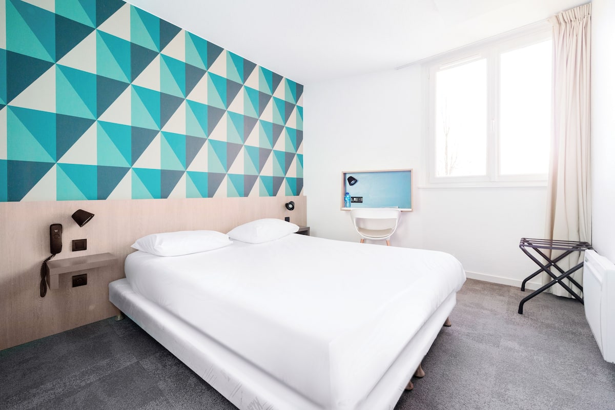 Ein Hotelzimmer mit einem Bett, einer Wand mit geometrischem Muster, einem Nachttisch mit Lampe und einem Vorhangfenster.