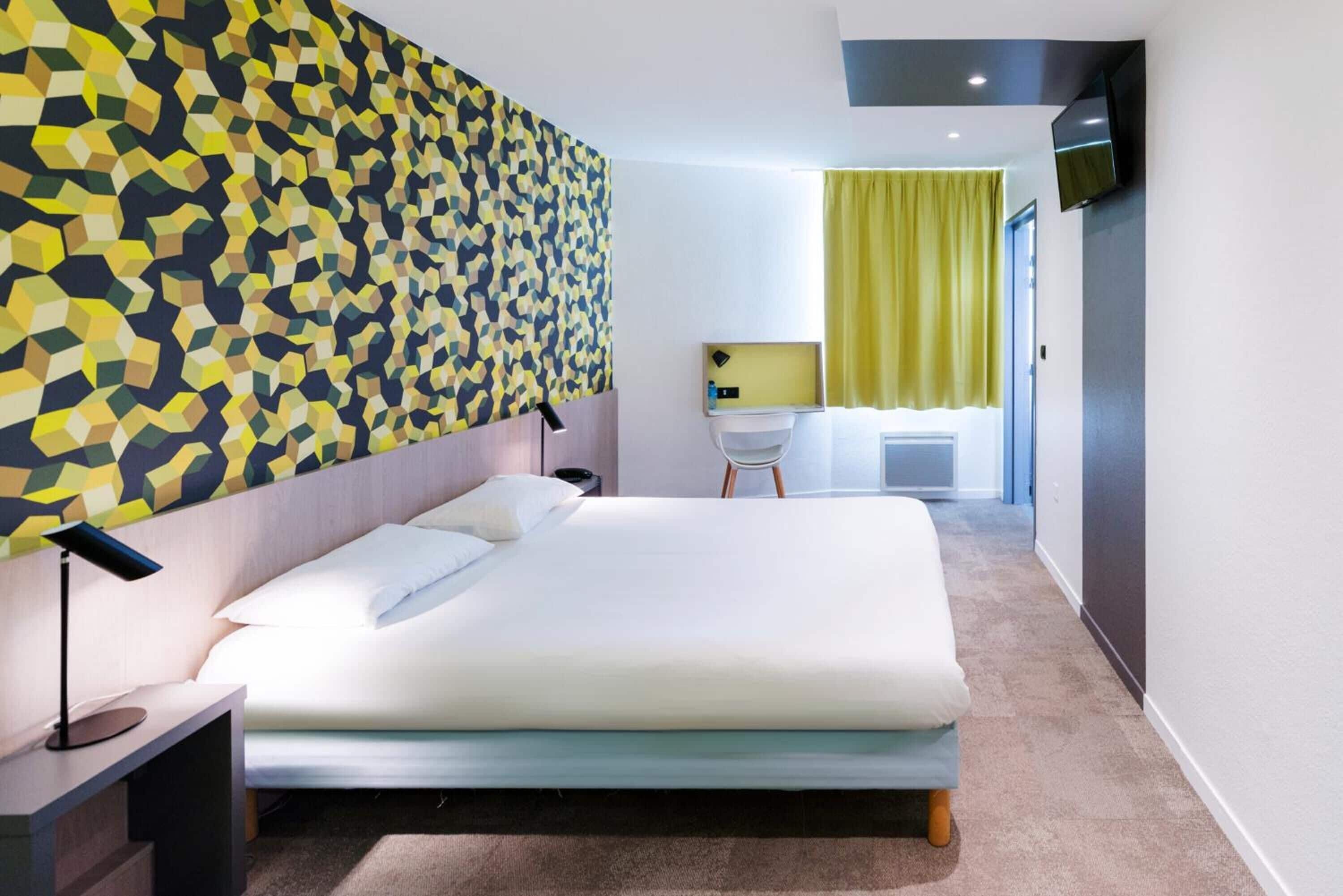 Een moderne hotelkamer met een wand met een geometrisch patroon, een bed met witte lakens, een bureau met stoel, en een aan de muur bevestigde televisie.