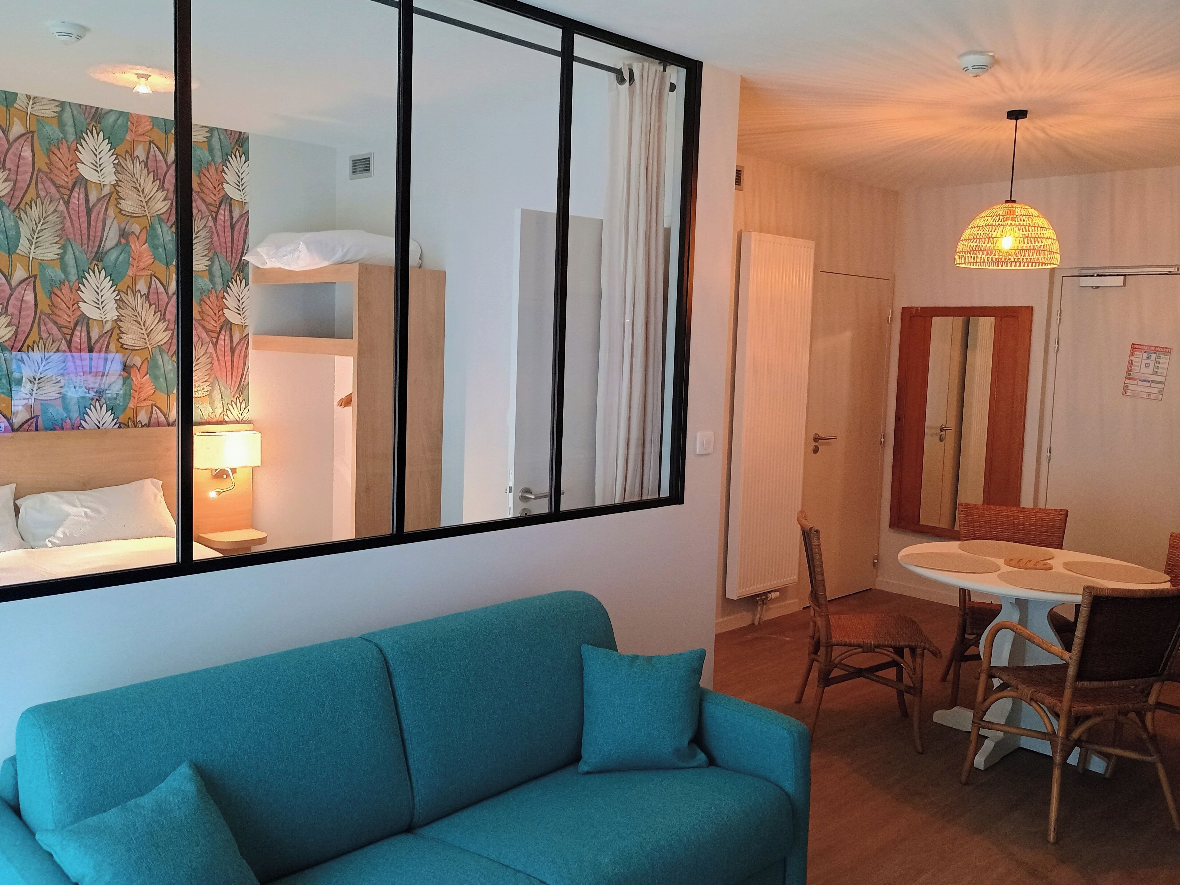 Une pièce avec un canapé de couleur turquoise, un coin repas avec des chaises en osier, un ventilateur de plafond et un mur orné d’une fresque à motifs de feuilles.