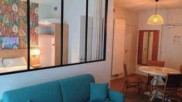 Une pièce avec un canapé de couleur turquoise, un coin repas avec des chaises en osier, un ventilateur de plafond et un mur orné d’une fresque à motifs de feuilles.