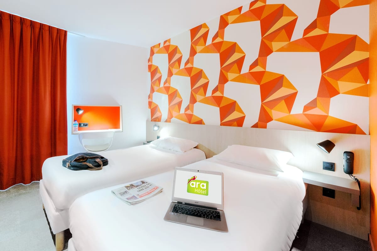 Ein Hotelzimmer mit zwei Betten, einem Schreibtisch mit einem Laptop, einer Schreibtischlampe, einem Wandfernseher und einem großen, geometrischen Wandbild.