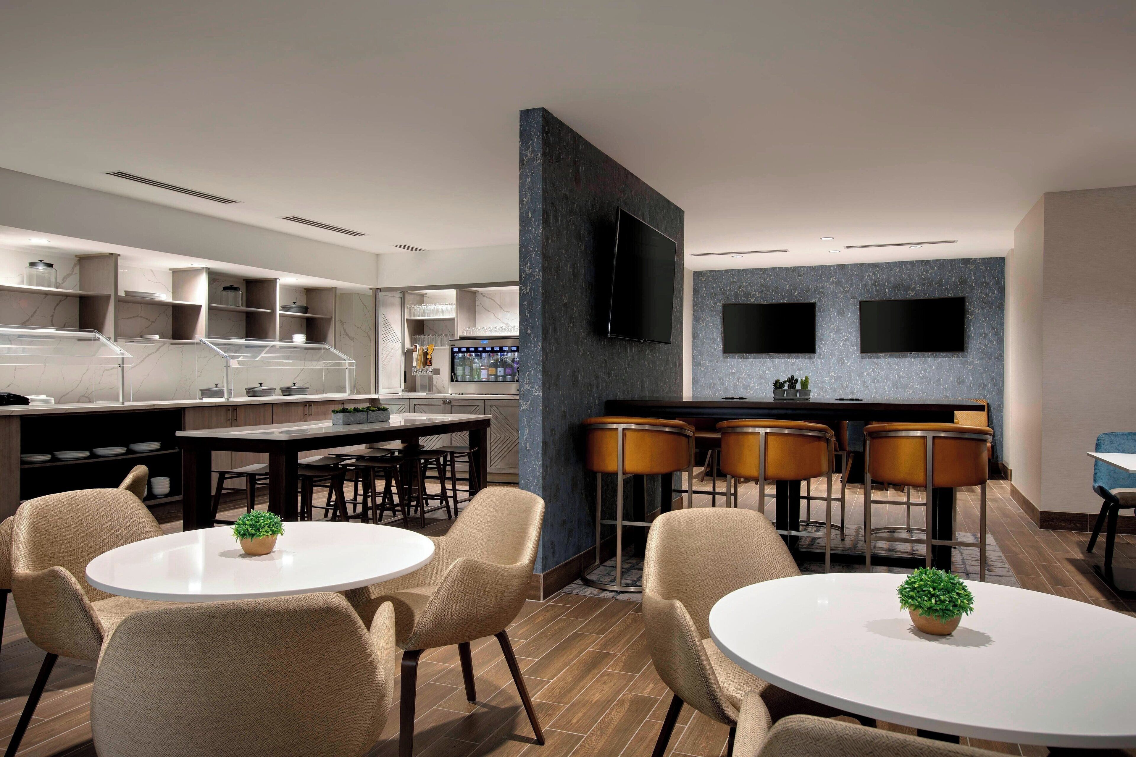A modern restaurant with round tables and chairs, a bar area with stools, and a kitchen in the background.
