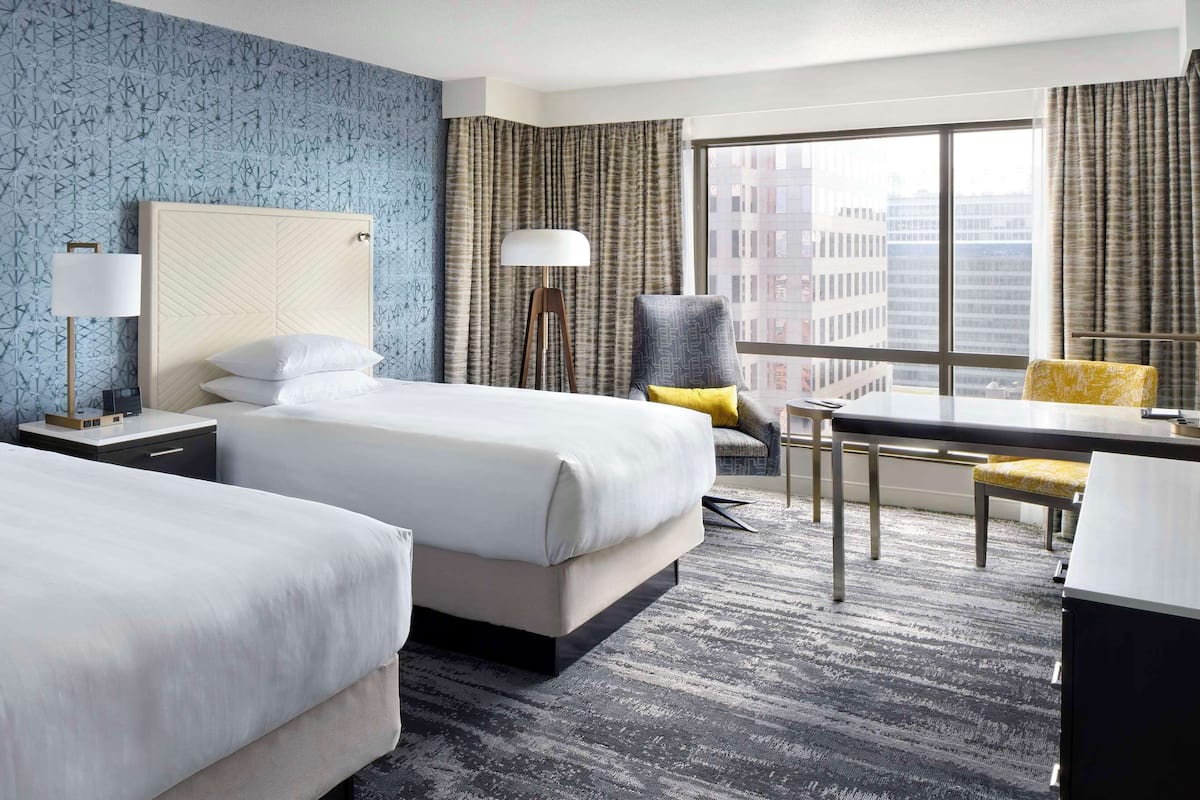 A hotel room with two beds, a desk, a chair, a TV, and a large window with curtains.