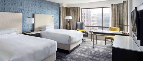 A hotel room with two beds, a desk, a chair, a TV, and a large window with curtains.