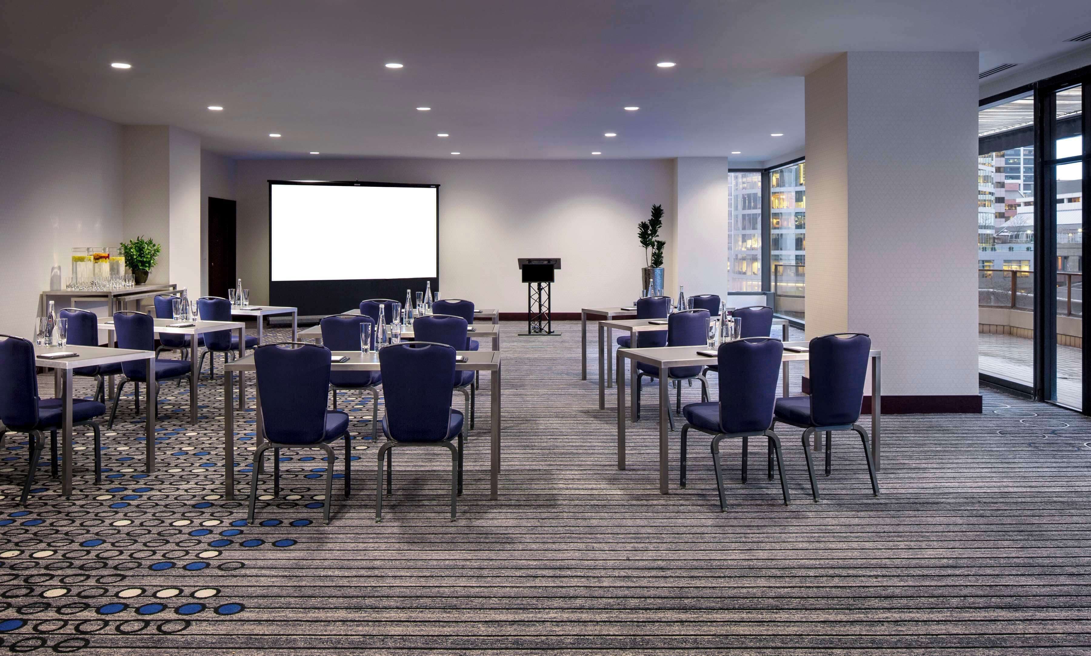 Un salón de conferencias con una pantalla grande, varias mesas y sillas dispuestas para reuniones.