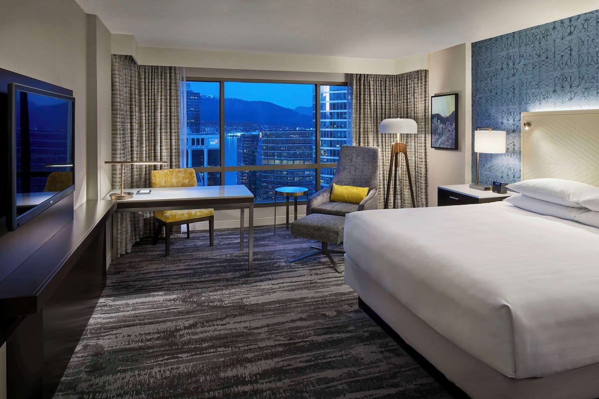 A hotel room with a large bed, a desk with a chair, a small table, and a view of a cityscape through large windows.