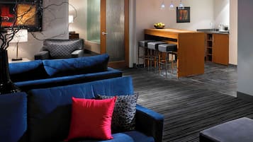 A modern living room with a blue sofa, a red pillow, and a black ottoman. There is a kitchen area with a bar and stools in the background.