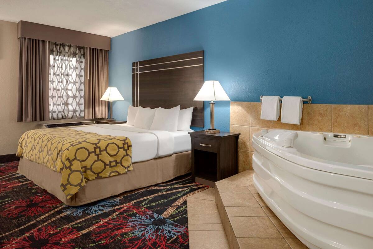 Habitación de hotel con cama, dos lámparas de noche, un ventanal con cortinas, un cabecero decorativo, una mesita de noche pequeña, una bañera blanca y una alfombra colorida.