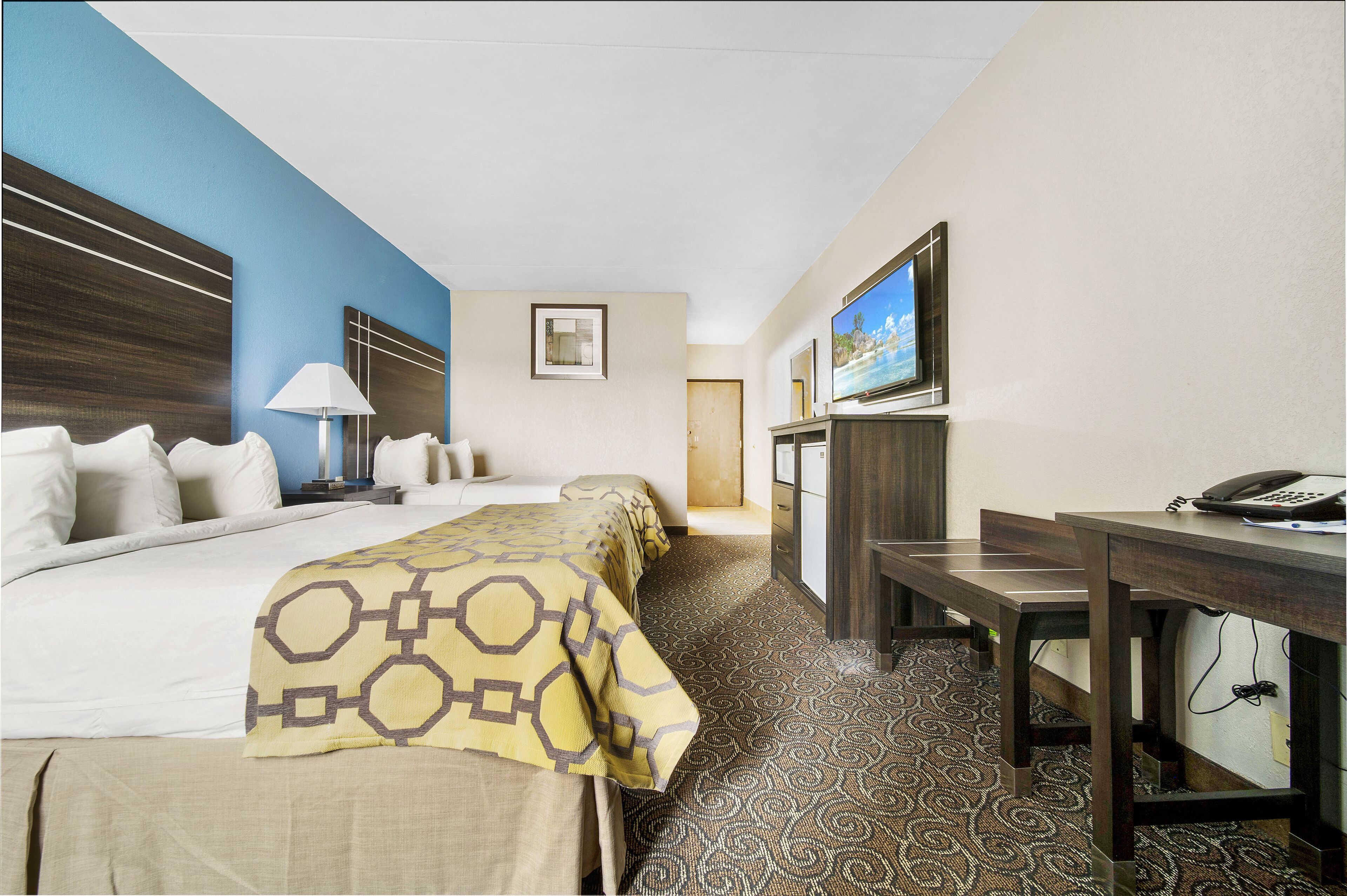 A hotel room with a bed, bedside table, lamp, TV, and a desk with a phone.