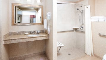 A bathroom with a granite countertop sink, a mirror with a decorative frame, a handheld hair dryer, and a walk-in shower with a glass door.
