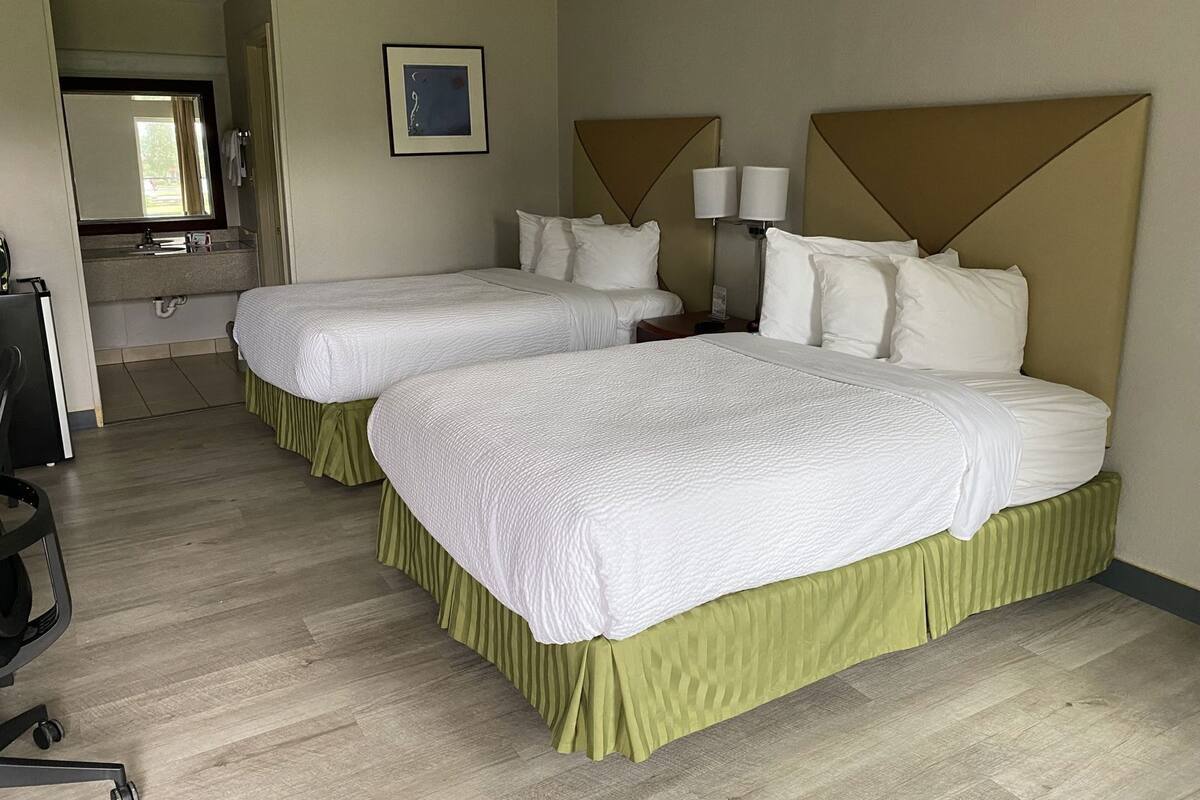 A hotel room with two beds, a desk, a chair, a mirror, a sink, and a framed picture on the wall.