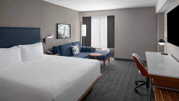 A hotel room with a large bed, a sofa, a small table, a desk, and a TV.