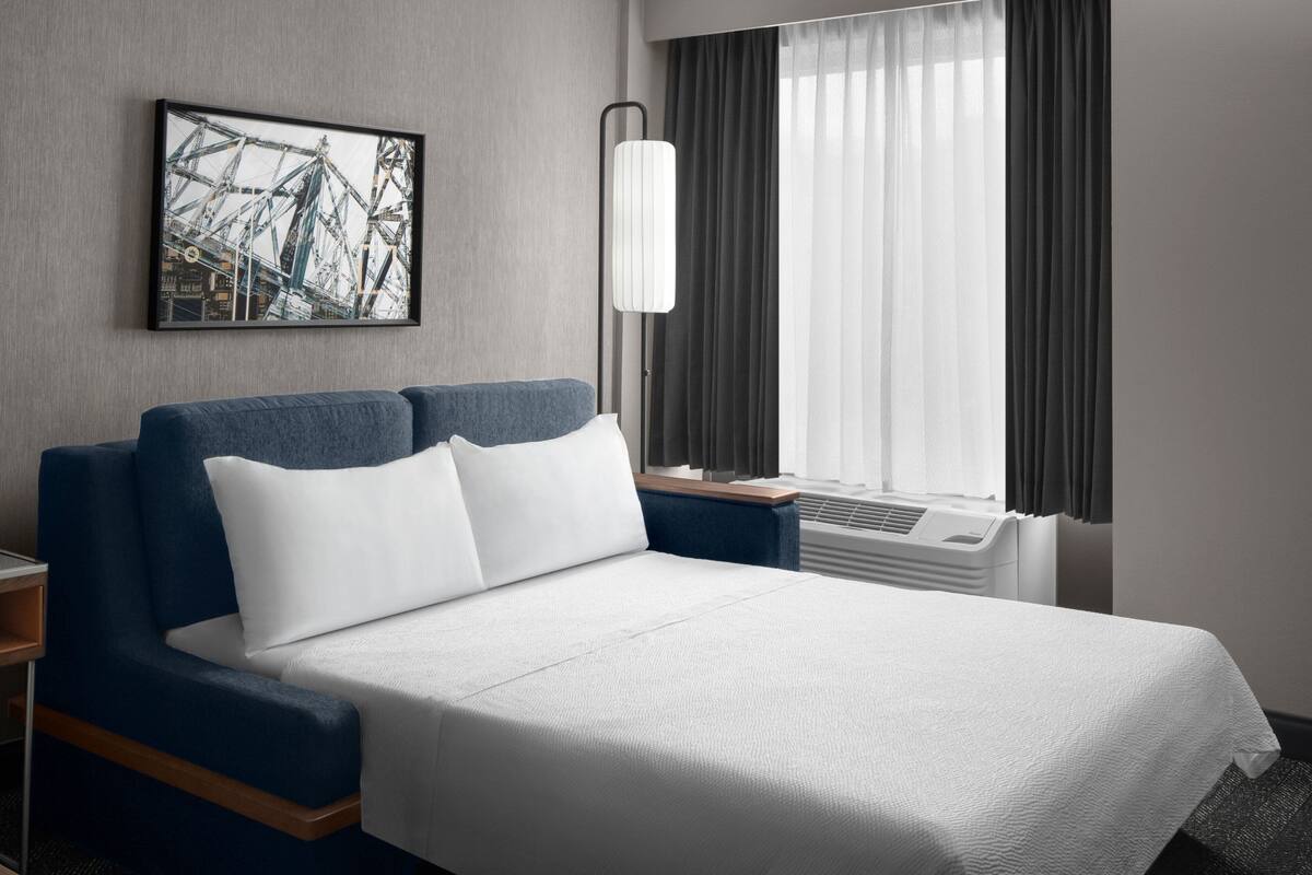 A hotel room with a bed, a nightstand, a lamp, a window with curtains, and a framed picture on the wall.