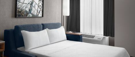 Una habitación de hotel con cama, mesita de noche, lámpara, ventana con cortinas e imagen en un marco en la pared.